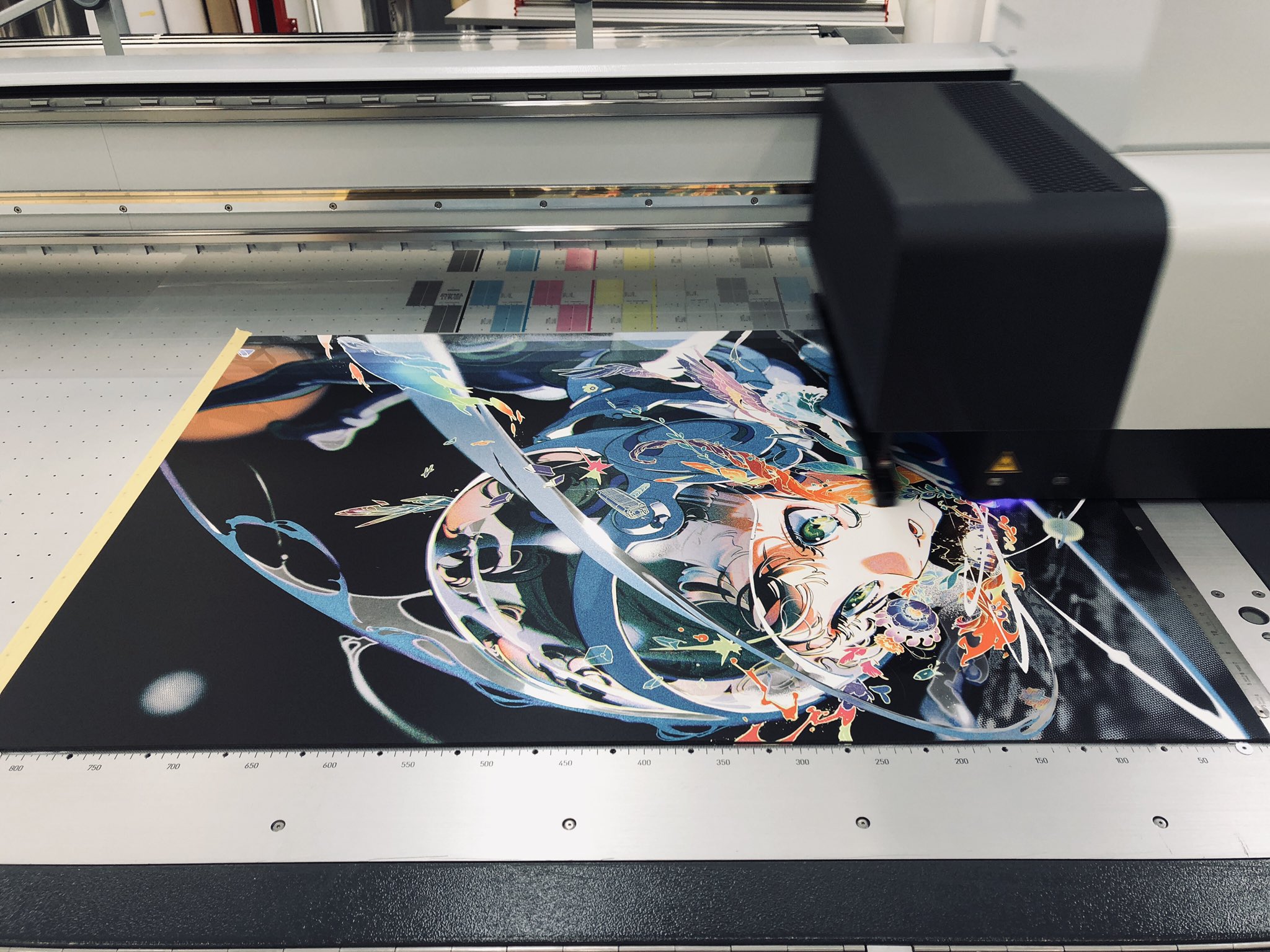 a large format printer is printing an animestyle graphic onto a piece of fabric. the print head is moving rapidly across the fabric, laying down ink in precise patterns. the artwork features a female character with flowing blue hair and a colorful, abstract background.  the printer is a large, industrial machine with a metal frame and a glass top. the room is brightly lit, and the printer is the focal point of the image.