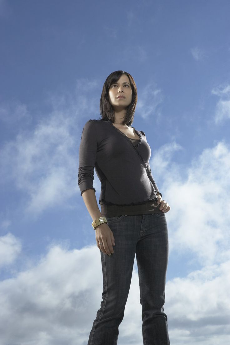 a woman in casual wear confidently stands with her hand on her hip against a bright blue sky filled with white clouds. the low camera angle emphasizes her powerful stance.