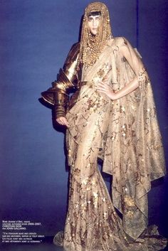 a woman in a gold sequined gown with an egyptian goddess theme poses for a fashion shoot. she is wearing an elaborate headpiece and necklace with gold accents, and she is wearing a lightcolored sheer gown that appears to be embroidered with gold threads. the woman is standing in a studio with a blue backdrop.
