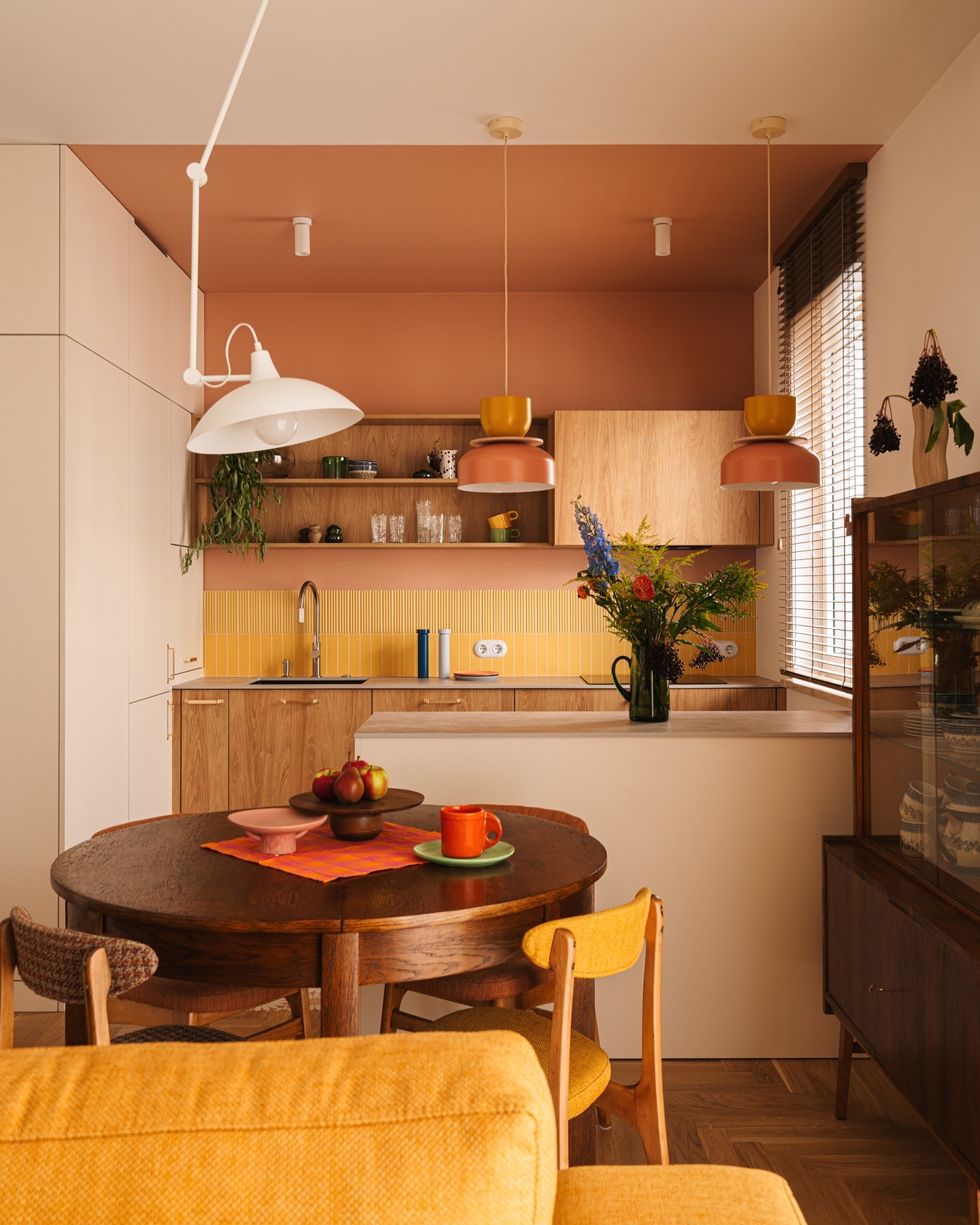 a kitchen and dining area with retroinspired decor. a round wooden dining table is in the foreground, surrounded by chairs with yellow and brown upholstery. a yellow sofa is partially visible in the lower part of the image. on the table there's a bowl of apples and an orange mug on a green plate. the kitchen features wooden cabinets, yellow tile backsplash, a sink, and pendant lights. the ceiling is painted in a terracotta color. a shelf above the countertop holds various items. the flooring is wooden parquet. a vintage cabinet with glass doors is visible on the right side. there are blinds on the window. a large white adjustable lamp hangs over the dining table. a vase of flowers sits on the counter next to the sink.