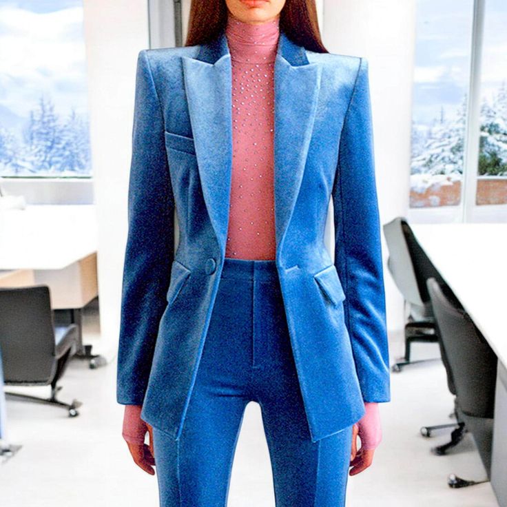 a woman in a blue velvet suit stands in an office setting. she is wearing a pink shirt underneath the blazer. the suit is tailored and fitted, and the woman is looking confident and stylish.