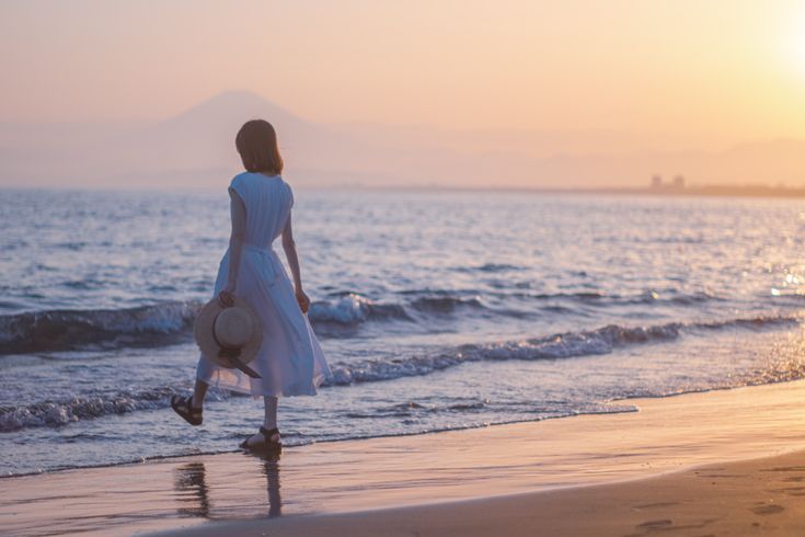 a woman in a white dress stands on a sandy beach at sunset, holding a hat and looking out at the sea. the water is calm with gentle waves, and a mountain is visible in the distance. the sky is a mix of orange, pink, and blue.