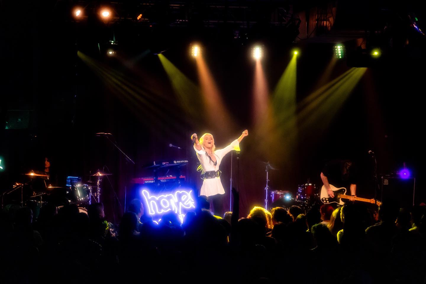 a musician is performing on stage in front of a crowd. she is a woman with blonde hair, wearing a white outfit and black belt, holding a microphone and raising her arms. she is standing in front of a keyboard and a neon sign that says "hope". behind her, there is a drum kit and another musician playing an electric guitar. the stage is lit with stage lights, and there are spotlights shining on the performer. the audience is visible in the foreground, with their heads and shoulders silhouetted against the bright stage.