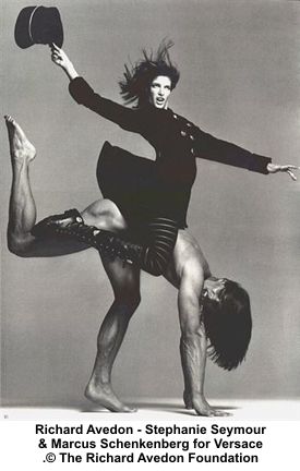 a black and white photograph of a fashion model wearing a black dress and hat, posing with a man in a studio setting. the model is standing on the man's back, her legs are raised and bent at the knee. the man's head is down, and he is looking at the ground. the image is a classic example of fashion photography from the 1990s, showcasing the glamour and style of the time.