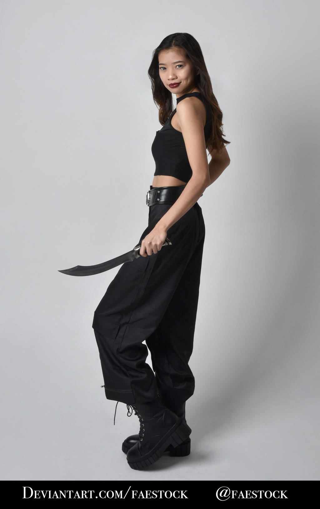 a fullbody, threequarter view of a young asian woman standing against a plain gray background. she is wearing a black crop top, black loosefitting pants, a black belt, and black combat boots. she is holding a black dagger in her right hand. she has long dark hair and a serious expression on her face. the image has a dark and edgy feel and could be used for stock photography or for character design.