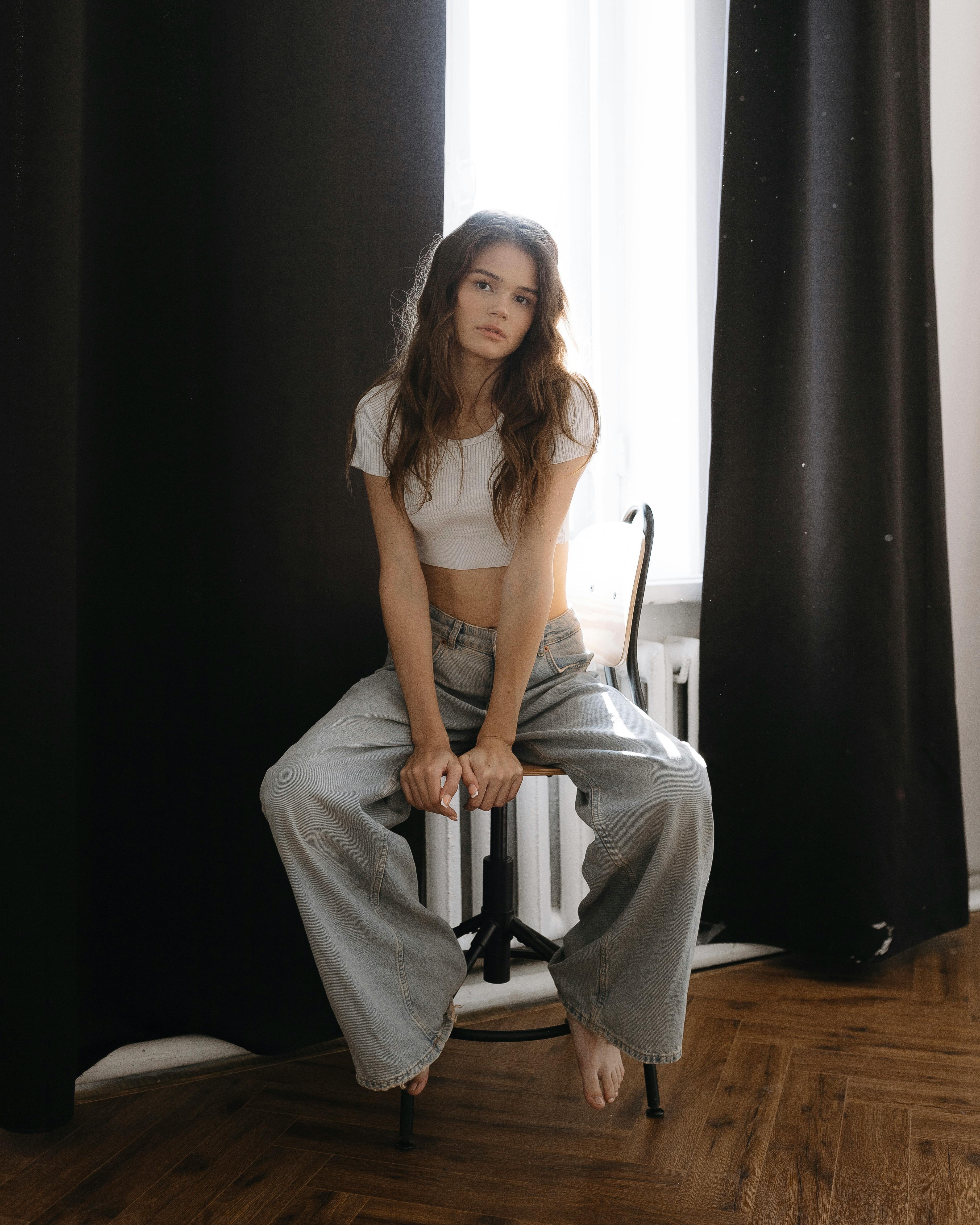 a young woman is sitting on a chair indoors. she is wearing a white crop top and baggy light blue jeans. she is barefoot. there are black and white curtains in the background. the floor is dark wood.