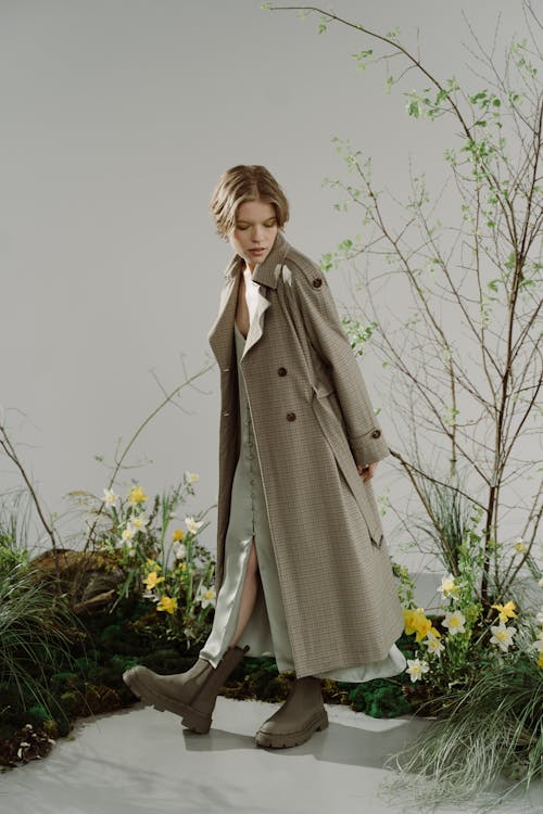 a full shot of a fashion model, a young woman with short brown hair, wearing a long, light brown checkered trench coat over a light sage green slip dress. she is wearing dark taupe chunky ankle boots. the model is posing outdoors amongst springtime plants and flowers, including daffodils. the overall tone of the image is soft and natural, with a focus on the clothing and the model's elegance. the background features a sparse, delicate arrangement of branches and greenery. the model appears to be walking, creating a sense of movement in the photo.
