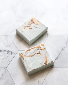 two square bars of soap, one on top of the other. the bars are white with a gray and gold marble swirl pattern. the background is a white marble surface.