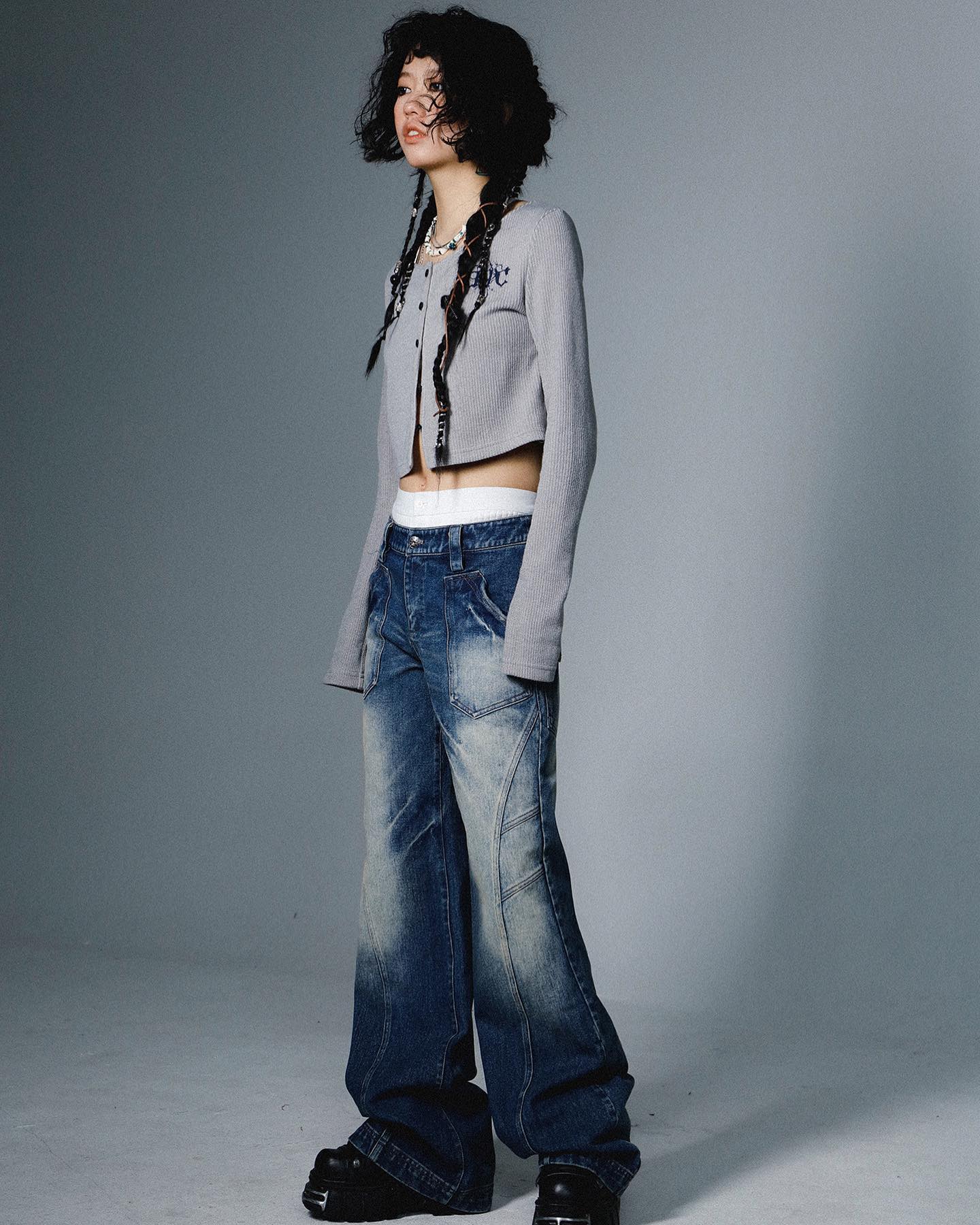 a fulllength studio shot of a young asian woman modeling a stylish outfit.  she has long dark hair styled in braids and is wearing a light gray cropped cardigan with long sleeves, a pair of lowrise distressed wideleg jeans, and chunky black platform shoes. her overall look is contemporary with a touch of grunge or y2k influence. the background is a simple, even gray that focuses the attention on the model and her clothing.