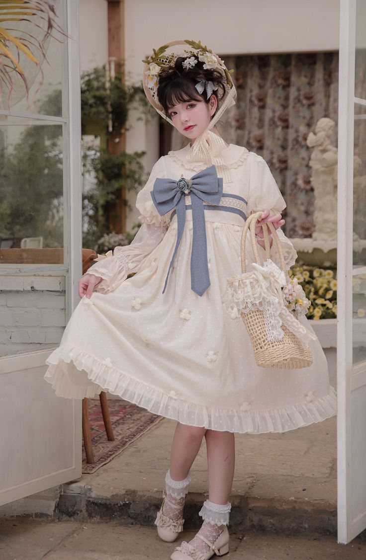 a young woman poses in a doorway while modeling a frilly, beige lolita fashion dress. the dress features floral appliques, puffy sleeves, and a large blue bow. she is also wearing a matching hat adorned with flowers, white socks, and mary jane shoes. she is holding a straw bag decorated with lace and flowers. the scene is brightly lit with natural light, suggesting an indoor setting like a greenhouse or sunroom.