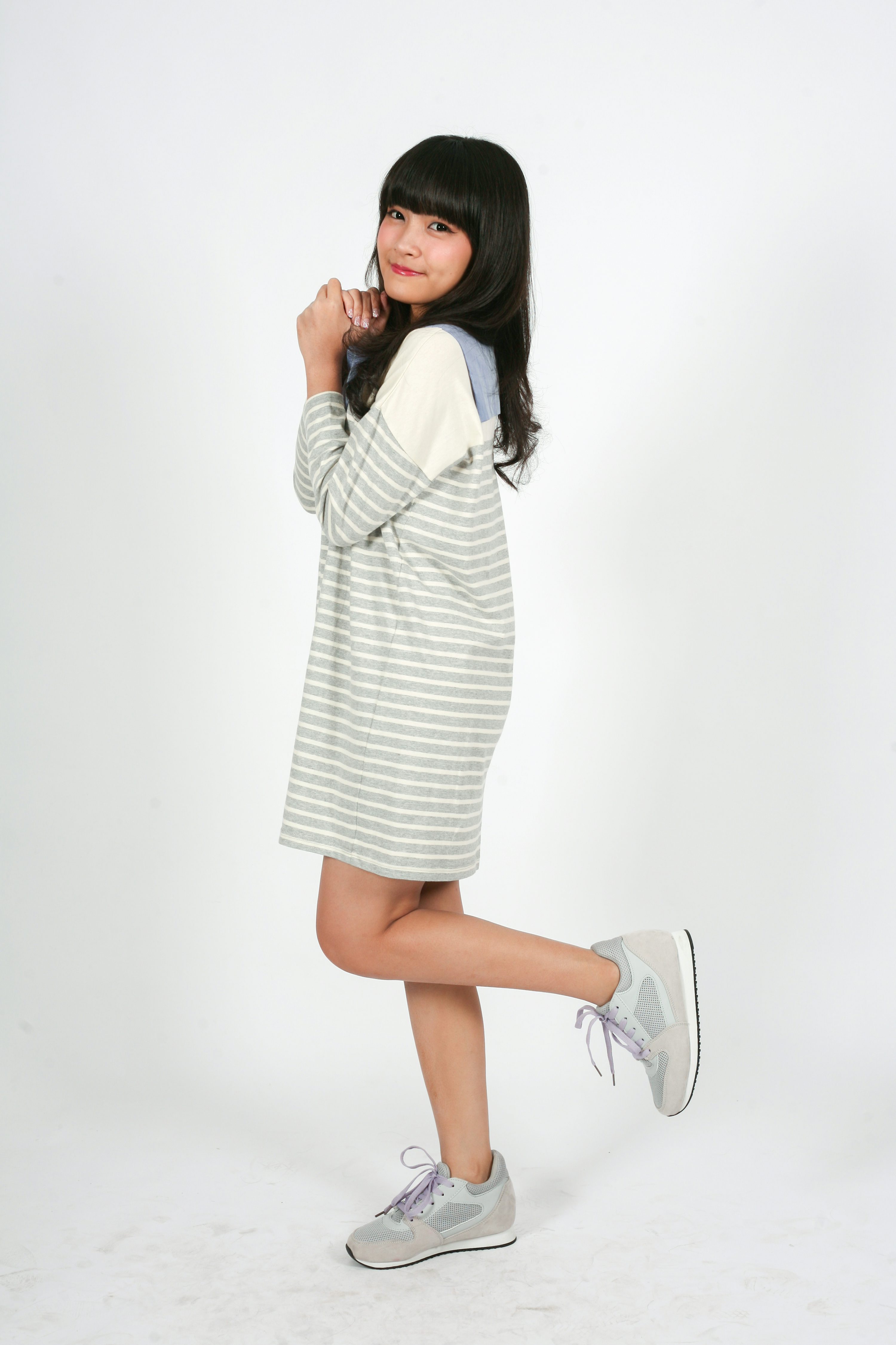 a young asian woman is standing in a studio against a white background. she is wearing a grey and white striped dress and grey sneakers. she is smiling and looking at the camera. her hands are clasped in front of her.
