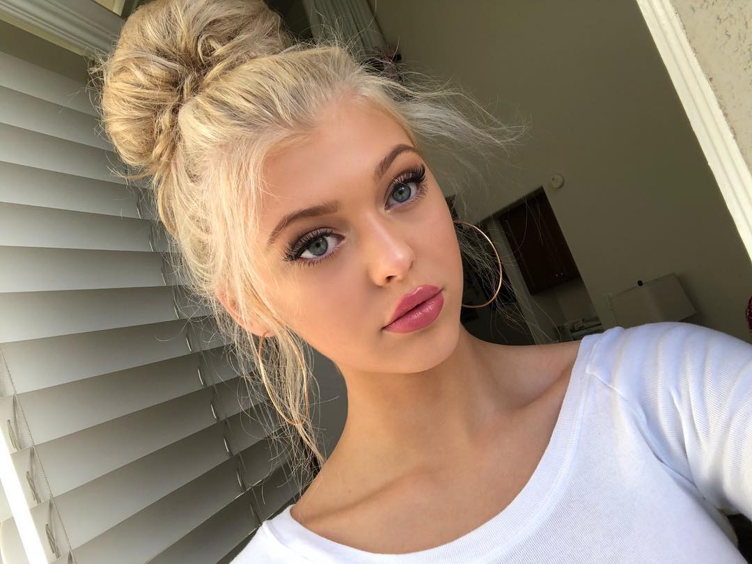 a closeup shot of a young woman with blonde hair styled in a high bun. she is wearing gold hoop earrings, a white shirt, and has on makeup including lipstick and enhanced eyelashes. she is standing in front of a window with closed blinds.