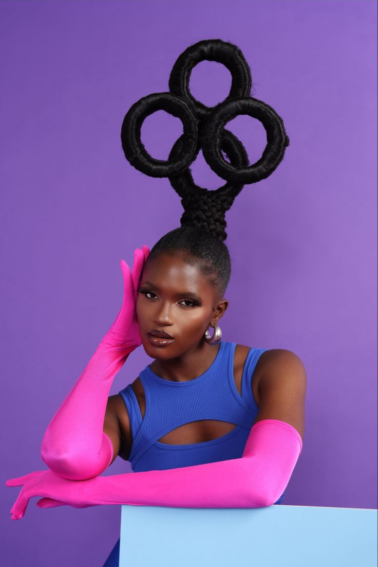 a young woman is wearing a bold,  blue crop top and pink gloves. she has a unique hairstyle that's styled into three circles. the woman has a beautiful, light complexion and is sitting with one hand on her face and the other resting on a blue surface. the background is purple.