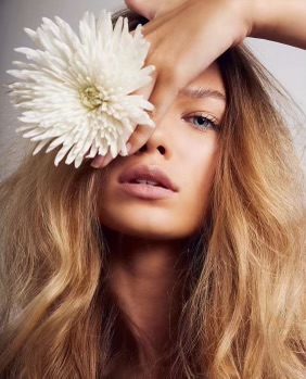 the image is a closeup portrait of a young woman with long, wavy blonde hair.  she is holding a large, white chrysanthemum flower up to her face, partially obscuring one eye. her makeup is light and natural, emphasizing her clear skin. the overall aesthetic is soft, elegant, and subtly sensual, focusing on the woman's natural beauty and healthylooking hair and skin. the background is a simple, light gray, keeping the focus on the model.
