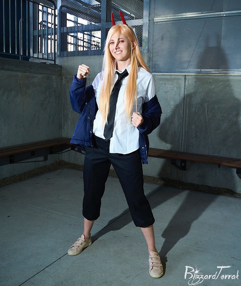 a fullbody shot of a young woman cosplaying a character, possibly from an anime.  she has long blonde hair with small red devil horns on her head. she is wearing a white shirt, black tie, dark cropped pants, and a navy blue jacket. her shoes are light beige sneakers. she's standing in a concrete area with metal railings and a bench visible in the background, striking a confident pose with one fist raised. her shadow is cast prominently on the ground behind her. the image has a slightly dark and moody atmosphere.  a watermark is visible in the bottom right corner.