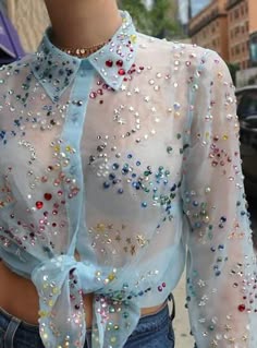 a close up view of a young woman wearing a light blue sheer shirt with silver, blue, and pink rhinestones. the shirt is tied at the waist and she is also wearing denim shorts.  the photo is taken from the chest up.