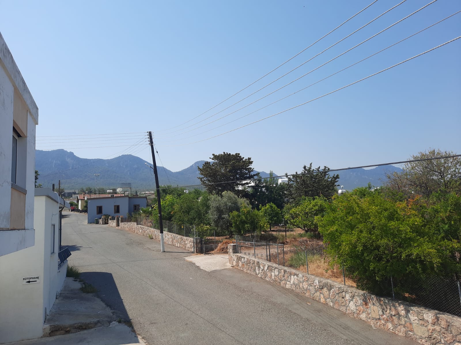 a daytime view of a road in a small town, lined with stone walls and trees, leading towards distant mountains under a clear blue sky. a leaning telephone pole stands along the road, and buildings can be seen on either side. an arrow sign points towards a library.
