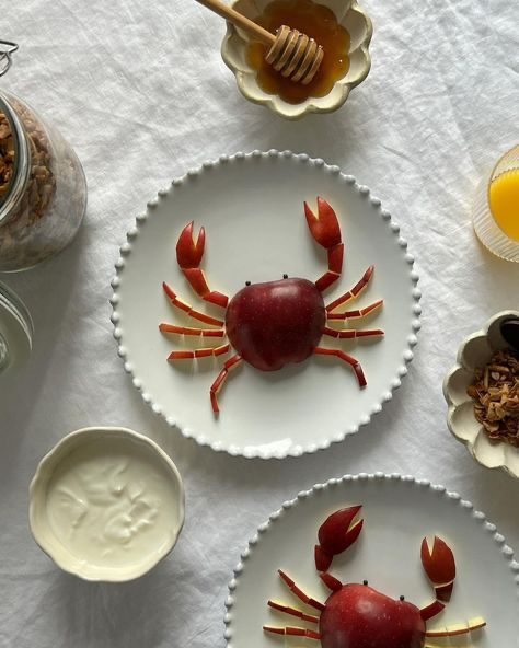the image shows a flatlay overhead shot of a meticulously arranged breakfast or snack.  two white plates feature apple crabs:  each crab is made from a whole apple as the body and apple slices arranged as legs and claws.  surrounding the plates are bowls of yogurt, granola, honey, and what seems to be some kind of topping in a small bowl. the overall style is minimalist and clean, with a focus on the artistic presentation of the fruit. the setting is a white tablecloth, creating a bright and airy feel.