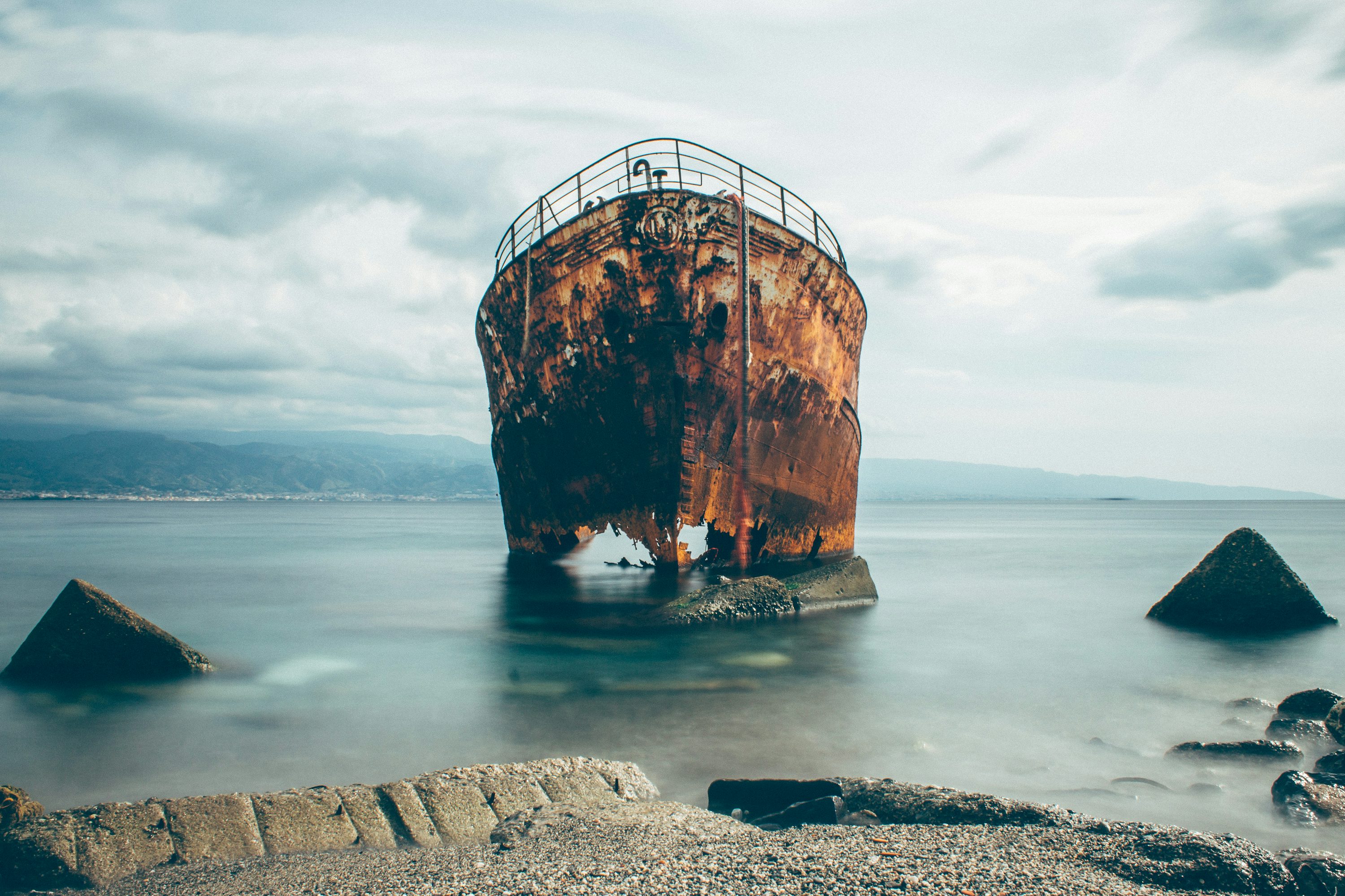 a rusty, weathered shipwreck stands partially submerged in calm ocean water. the bow of the ship is broken and jagged. large rocks surround the wreck, possibly acting as a breakwater. the background features a mountain range with a thin layer of clouds overhead. the overall color palette is muted and cool, suggesting a cloudy or overcast day.