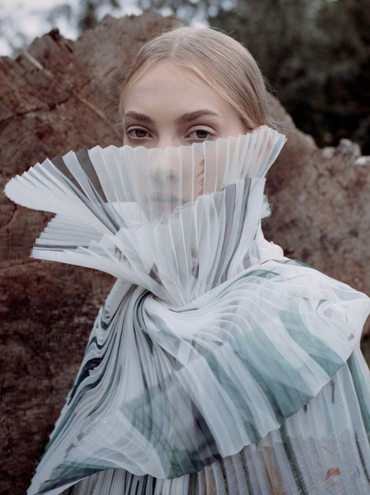 a woman with blonde hair stands outdoors with a large tree trunk behind her. she is wearing a garment made of pleated, lightcolored fabric that covers the lower half of her face, leaving only her eyes visible.