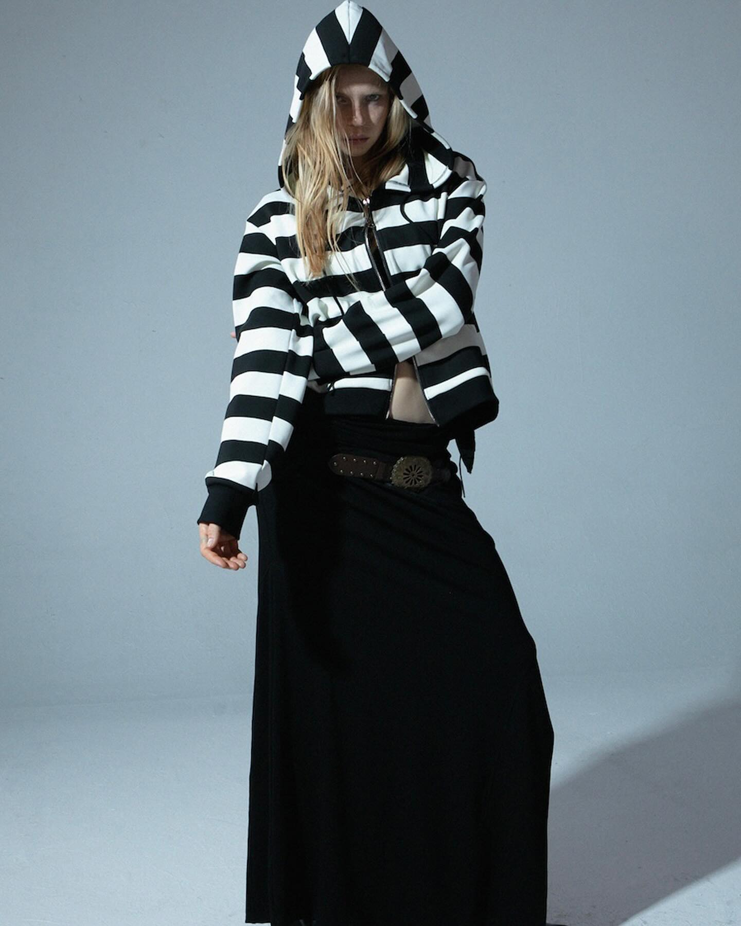 a full shot of a fashion model posing in a studio. she is wearing a black and white striped hooded jacket and a long black skirt. her blonde hair is visible beneath the hood. she is wearing a darkcolored belt with a circular buckle.  the background is a plain grey.