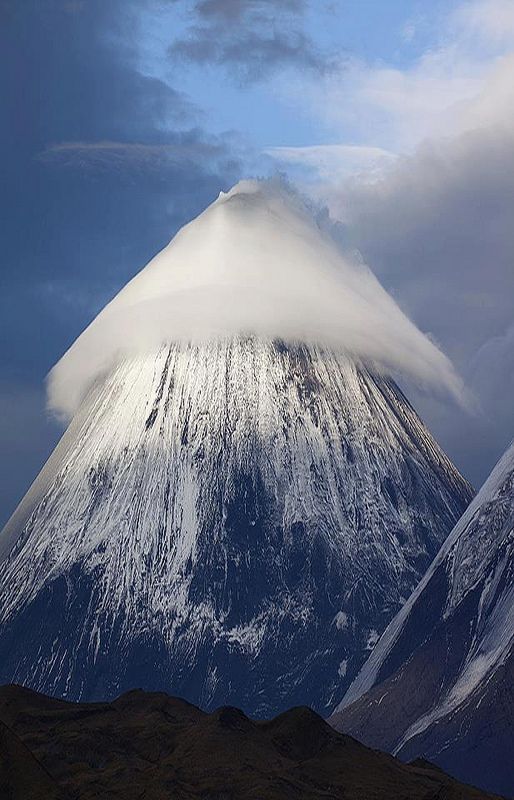 a majestic mountain peak stands tall against a backdrop of a clear blue sky and fluffy white clouds. the mountain is covered in snow, and its slopes are steep and rugged. the clouds create a dramatic effect, as if they are swirling around the peak. the scene is one of natural beauty and aweinspiring power.