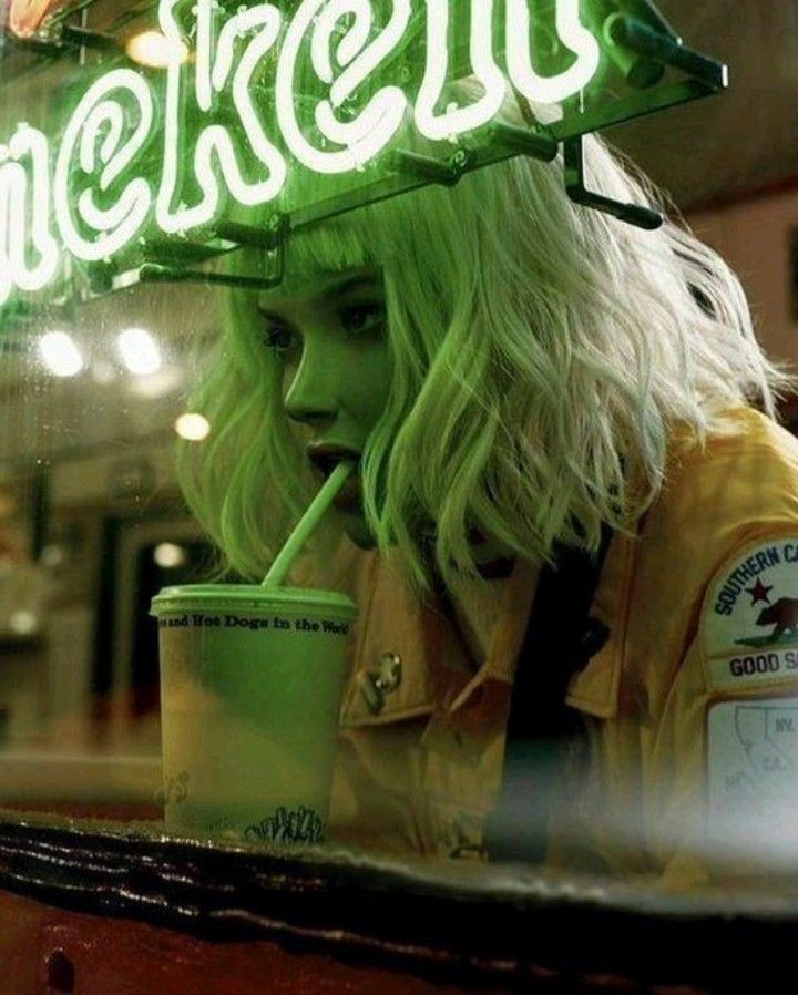 a young woman with bright blonde hair is sitting in a cafe. she is wearing a yellow jacket and looking down at a green cup. there is a green neon sign above her head. the image has a vintage feel.