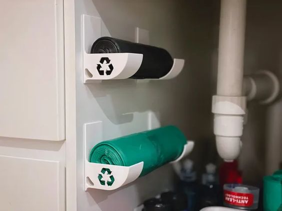 a white cabinet with two white garbage bag organizers mounted underneath the sink, one holding black garbage bags and the other green trash bags.  the organizers have a recycling symbol on them.