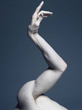 a closeup, partial view of a person's arm, extending gracefully upwards against a muted blue background. the arm is muscular, with soft lighting highlighting its contours and creating a sense of depth. the pose suggests a sense of elegance and fluidity, reminiscent of classical sculpture or dance.