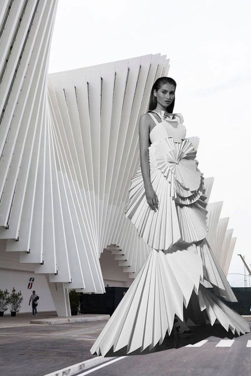 a fashion model in a white pleated dress poses in front of a modern, white architectural building. the dress is made of many layers of fabric that are folded and pleated to create a dramatic and flowing effect. the building is also made of white, folded panels, which creates a sense of harmony and unity between the model and her surroundings. the image is minimalist and stylish, with a strong sense of design.