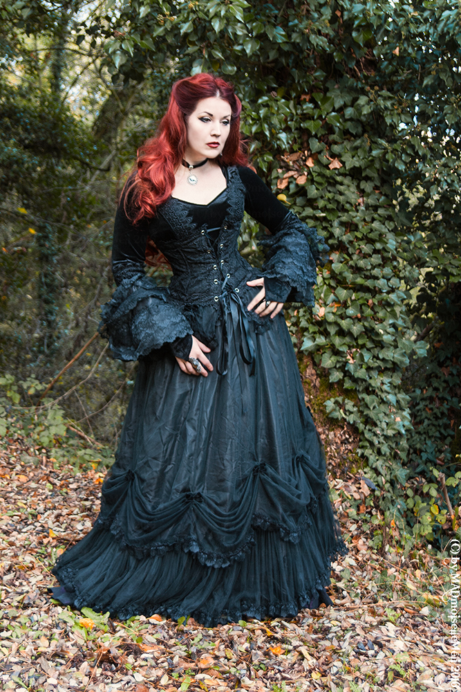 a woman dressed in gothic fashion stands in a forest. she has red hair and is wearing a black corset dress with long sleeves and a ruffled skirt. she is also wearing a choker necklace and a ring. the background is a forest with fallen leaves on the ground.