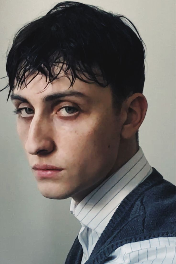 a portrait of a young man wearing a white shirt and a blue vest. he has short, dark hair and a serious expression. the photo was taken indoors and the background is simple and neutral.