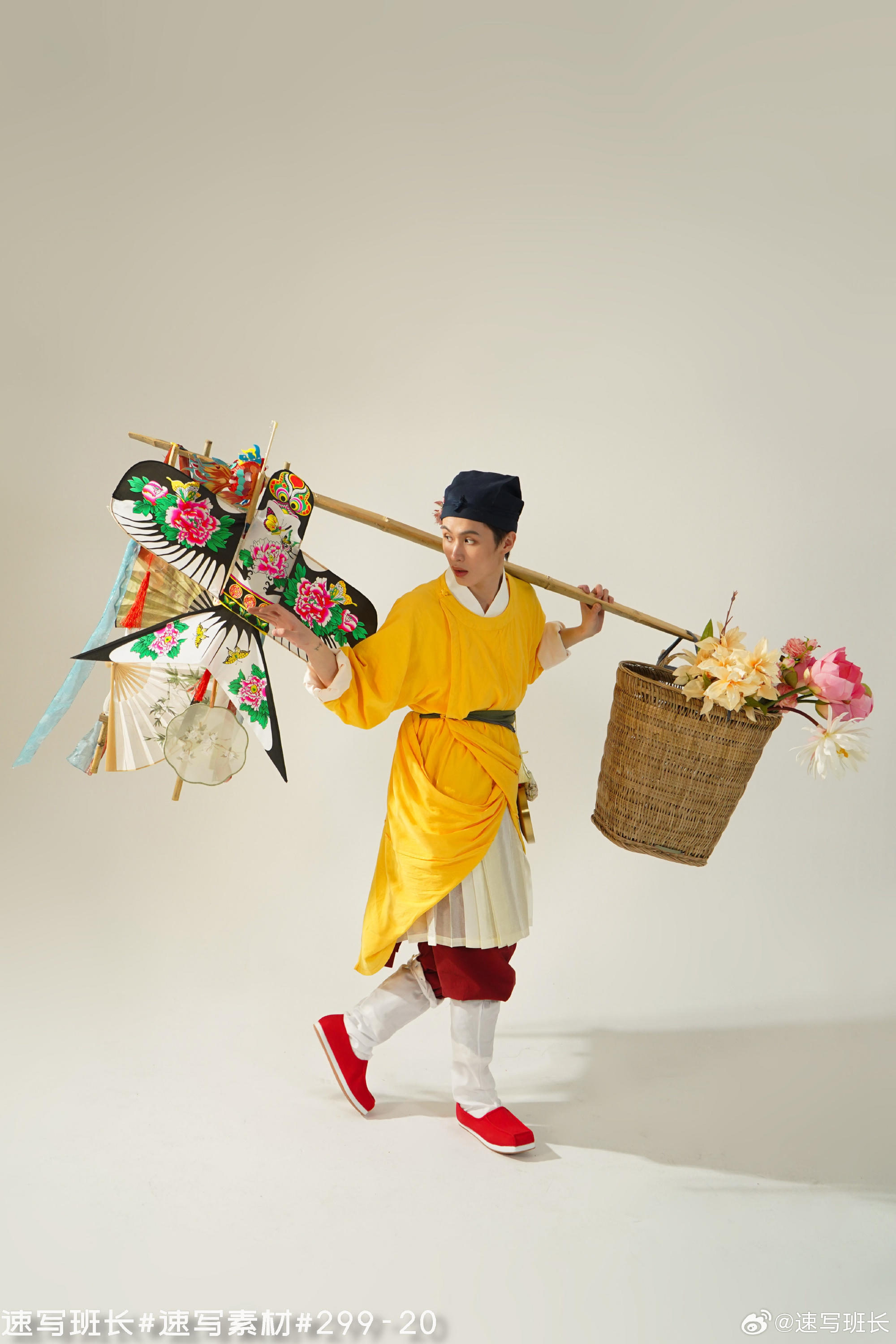 a man dressed in traditional chinese clothing is walking. he is carrying a large, colorful kite on his shoulder, as well as a basket of pink and white flowers. the image appears to be a studio shot with bright, even lighting.  the man is wearing red shoes, a yellow robe, white pants, and a blue hat.