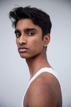 the image is a portrait of a young man with dark hair and brown eyes. he is wearing a white tank top and has a serious expression on his face. the lighting is soft and the background is a light gray. the perspective is a side view.