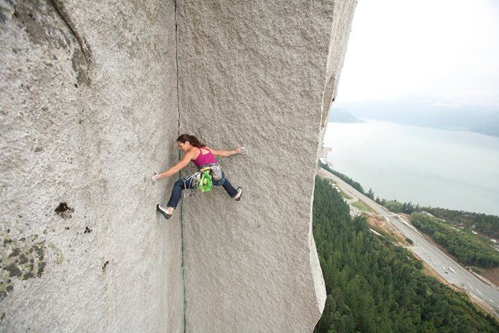a female rock climber scales a steep, sheer rock face with a lake and forested mountains in the background. she is positioned horizontally with her left hand reaching for the next hold and her right leg extended for balance. she's wearing a pink tank top, blue climbing pants, and has climbing gear attached to her harness.