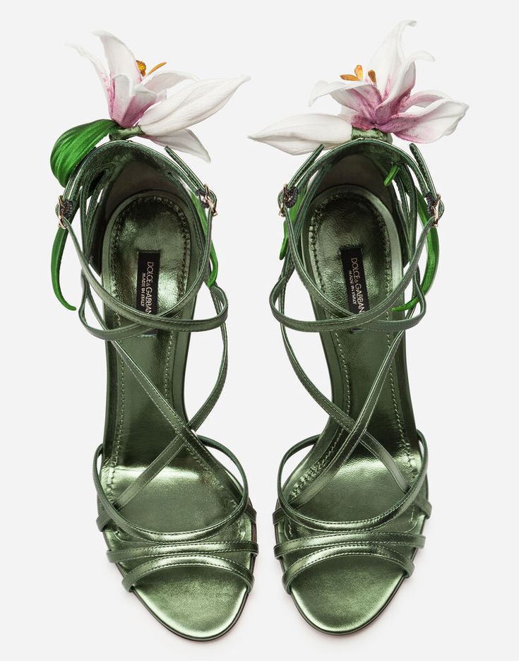 the image shows a pair of women's dolce & gabbana highheeled sandals.  the sandals are a sage green metallic leather with intricate crisscross straps.  each sandal is adorned with a large, realisticlooking white and pink lily flower. the shoes are presented from an overhead perspective on a white background, showcasing their detail and craftsmanship.