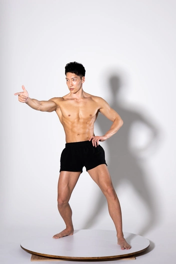 a muscular man with a toned physique is posing in a studio against a white background. he is wearing black shorts and is striking a dynamic pose, pointing with one arm and the other hand resting on his hip. his body is clearly defined, showcasing his abs, chest, arms, and legs. the overall image conveys fitness, strength, and athleticism.  he's standing on a round, white platform.