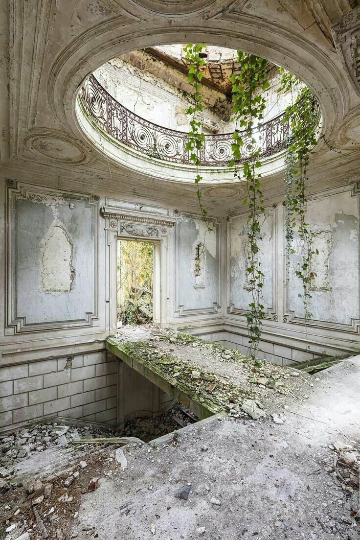 the image shows an abandoned and decaying interior of a building. the room features classical architectural elements such as ornate wall panels, a circular opening in the ceiling with a decorative balcony, and an arched doorway leading to the outside. the walls are crumbling, the paint is peeling, and there is significant decay and debris throughout the room. ivy grows down from the ceiling opening, and the overall scene conveys a sense of ruin and abandonment.