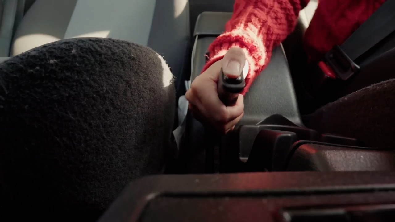 a closeup shot from the perspective of a driver pulling the handbrake of a car. the driver is wearing a red sweater. the interior of the car is dark and lit by sunlight coming through the windows.