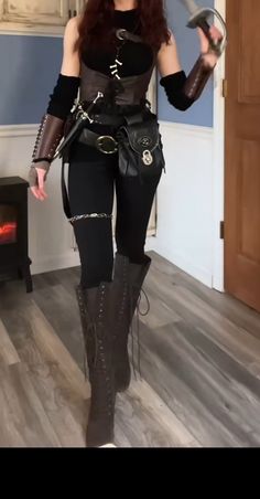 a woman is posing indoors, showcasing a cosplay outfit. she is wearing a black corset over a black shirt, black pants with chain details, tall laceup boots, and arm gauntlets. the outfit also features a belt with multiple pouches and sheaths for daggers. she holds a prop sword in her right hand. the image suggests a fantasy warrior or rogue character. the room features wooden flooring and a whitepainted wooden door. a fireplace is partially visible on the left side of the image.