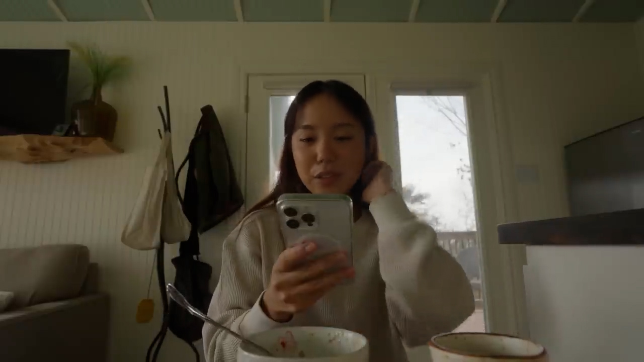 a young woman sits at a small table in a tiny house. she is looking at her phone and holding it in her right hand while seeming to tuck her hair behind her ear with her left. she is lit by natural light coming from a window behind her. it appears she is eating breakfast as there is a bowl with remnants of food in front of her. the image has a cozy and personal atmosphere, likely a selfie from a vlog.