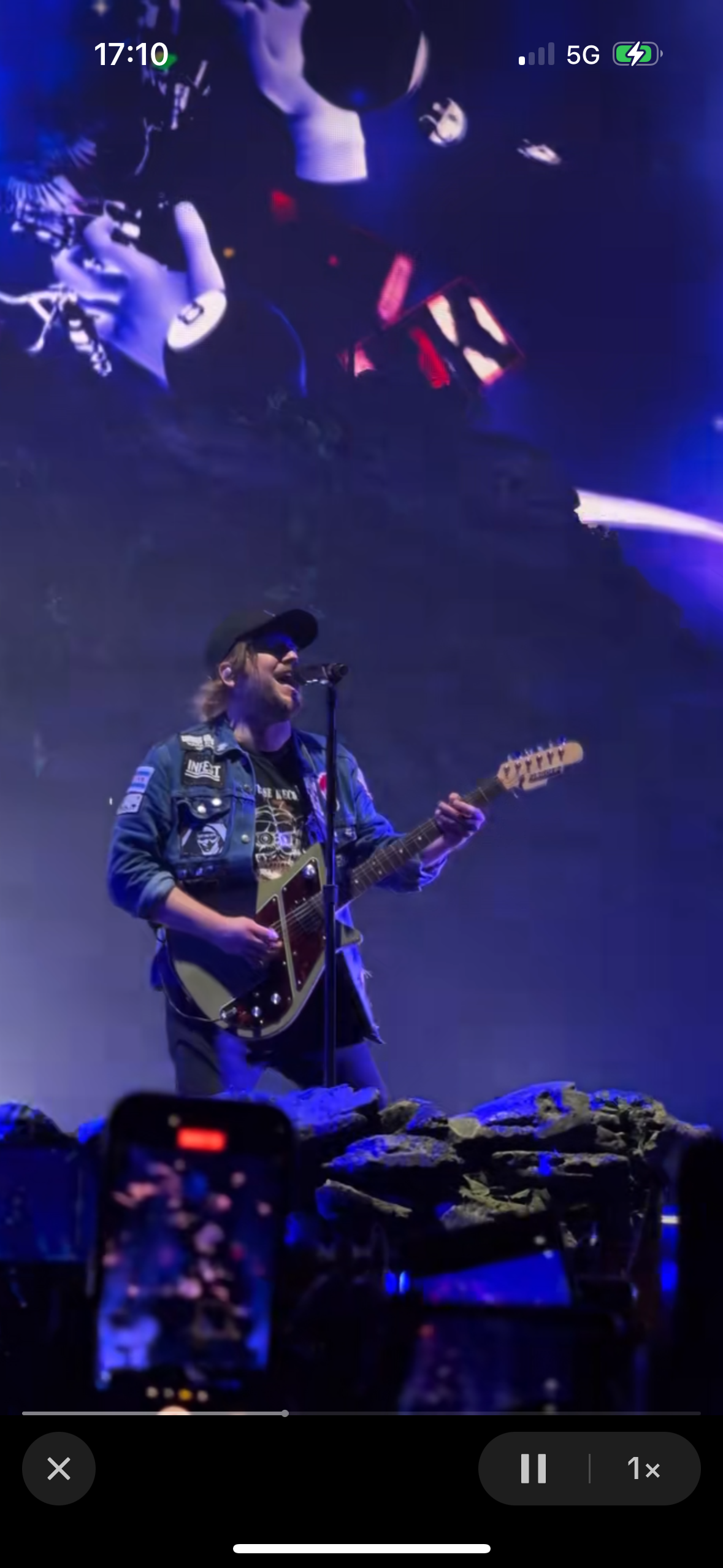 a male musician performs live on stage, singing into a microphone and playing a blue electric guitar. he is wearing a blue and black jacket and a black tshirt. the stage is dark and the crowd is blurred out in the background. the stage is lit with blue and purple spotlights. the musician is moving dynamically and looks engrossed in the performance. the overall atmosphere is vibrant and energetic.