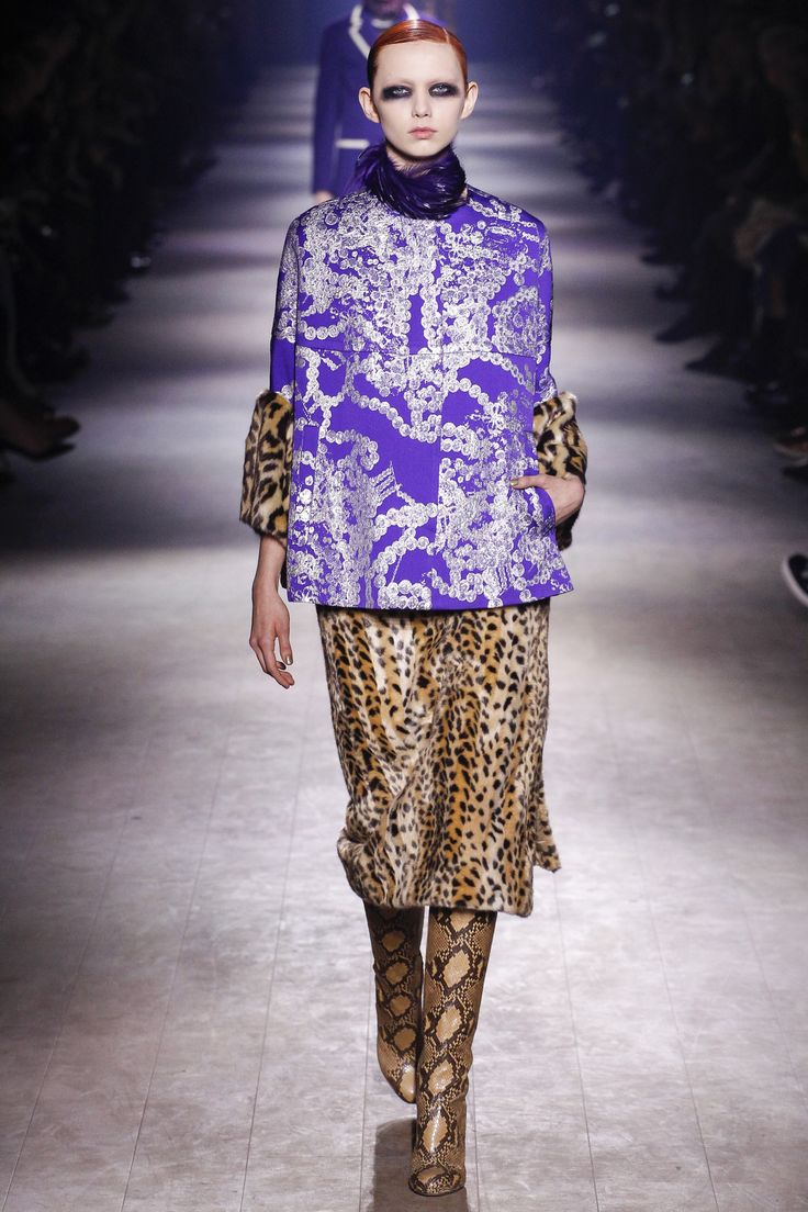 a model walks down a runway wearing a purple coat with a silver chain pattern, a leopard print skirt, and snakeskin boots. the overall style is high fashion and elegant, suitable for a fall or winter collection. the runway setting adds to the context of the image. the model's makeup and hair are also stylish and complement the outfit.