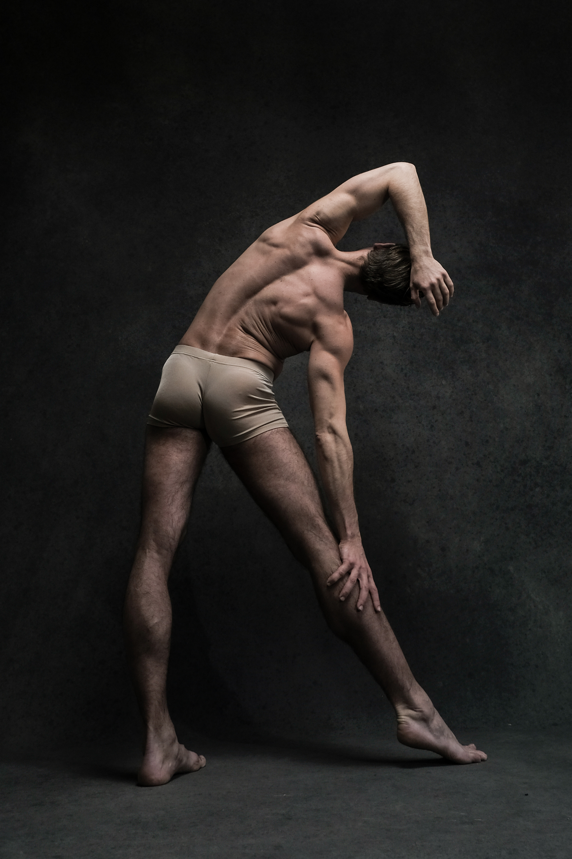 a male dancer is shown from the back, stretching in a dramatic pose against a dark studio background. the lighting emphasizes the musculature of his back, legs, and arms. the dancer's pose suggests both strength and flexibility.