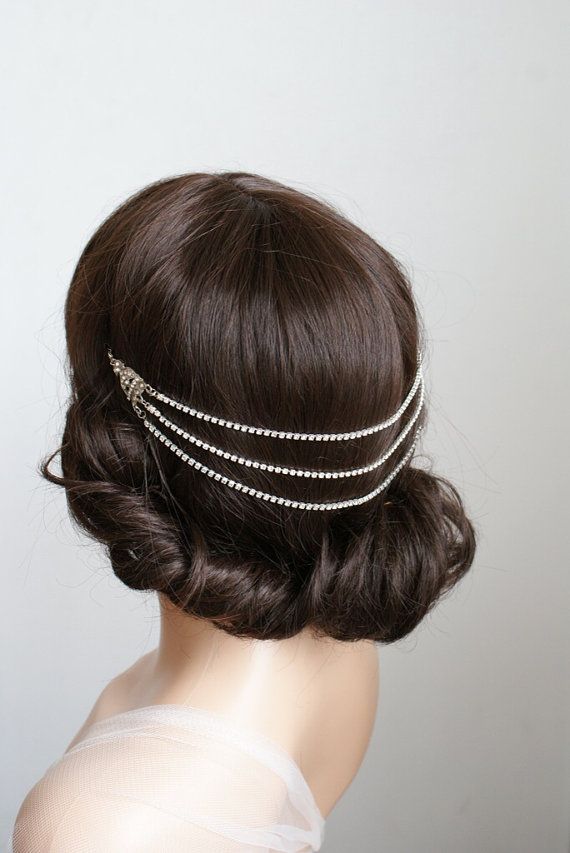 a woman with dark brown hair is wearing a  dainty crystal crown headband that is a chain of small stones with a few larger ones.  the headband has three rows of stones that are evenly spaced. her hair is pulled back in a simple style, creating a sophisticated look. her hair is smooth and even. the woman is wearing a white, sheer dress, the fabric of which looks like tulle or organza,  and is likely bridal.   the background is a plain white wall. the overall look is elegant and timeless.