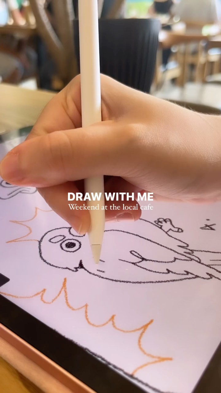 the image shows a closeup of a person's hand holding an apple pencil and drawing a simple sketch of a bird on an ipad. the drawing is done in a loose, casual style, with simple lines and shapes. the background is slightly blurred, revealing a café setting.  text overlay reads "draw with me, weekend at the local cafe". the overall feel is relaxed and informal.