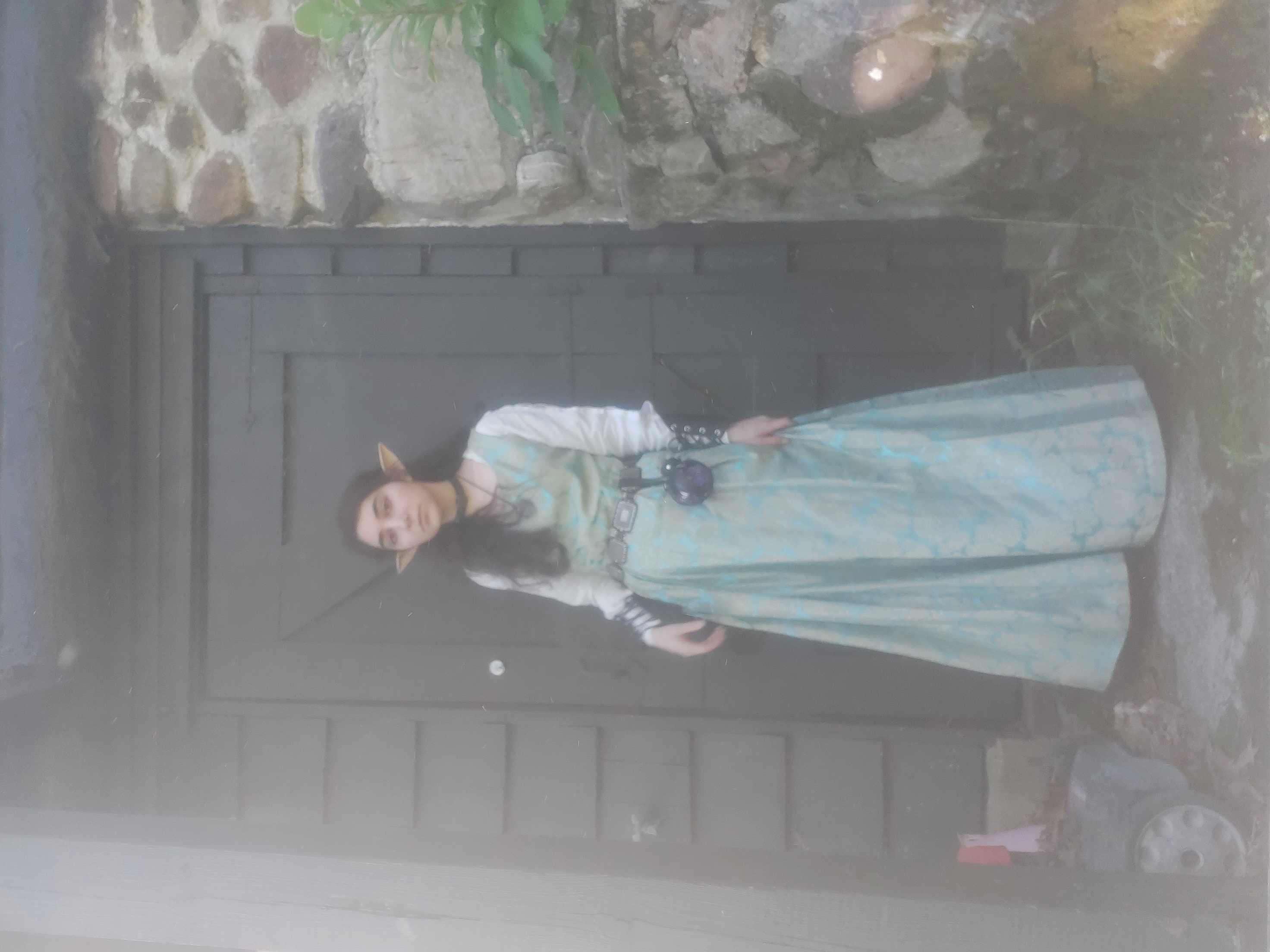 the image shows a young woman dressed in an elf costume. she's wearing a long, teal dress with a somewhat medieval or fantasy style, and has pointed elf ears. she stands in front of a dark brown wooden door set into a stone wall.  the scene has a slightly hazy or misty quality. the overall ambiance is one of fantasy and possibly cosplay.