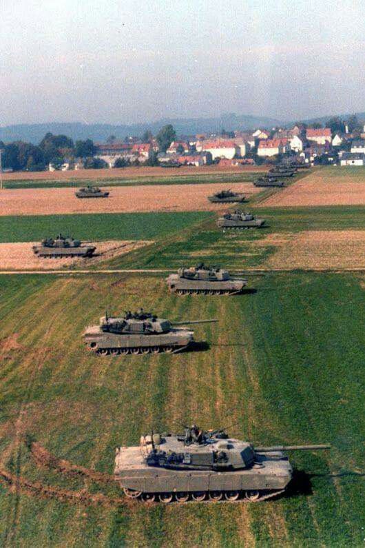 the image shows a long column of m1 abrams tanks moving across a vast, flat, green and brown farmland. the tanks are spaced apart and follow a roughly linear path across the fields. in the background, a small village or town is visible, suggesting a rural setting. the overall impression is that of a military exercise or maneuver, possibly during the cold war era given the era of the m1 abrams. the aerial perspective provides a broad view of the column and its surroundings. the image is slightly grainy, suggesting it's an older photograph.