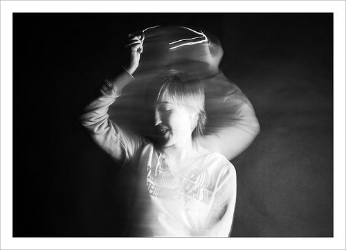 a black and white portrait of a young woman with a dark background and a light spotlight. her hair is dark and long, and she is wearing a white shirt that is blurred from motion. she appears to be in thought or dreaming, with a soft smile on her lips.