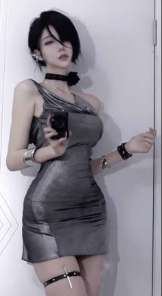 a woman with black hair and a silver dress is standing in a white studio. she's looking at the camera, with a silver choker and a black leather strap on her thigh.